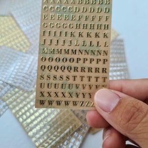 May include: A rectangular sheet of gold-coloured adhesive alphabet stickers featuring uppercase letters arranged in rows. The sheet contains multiple instances of each letter from A to Z, presented in a serif font style on a clear backing.