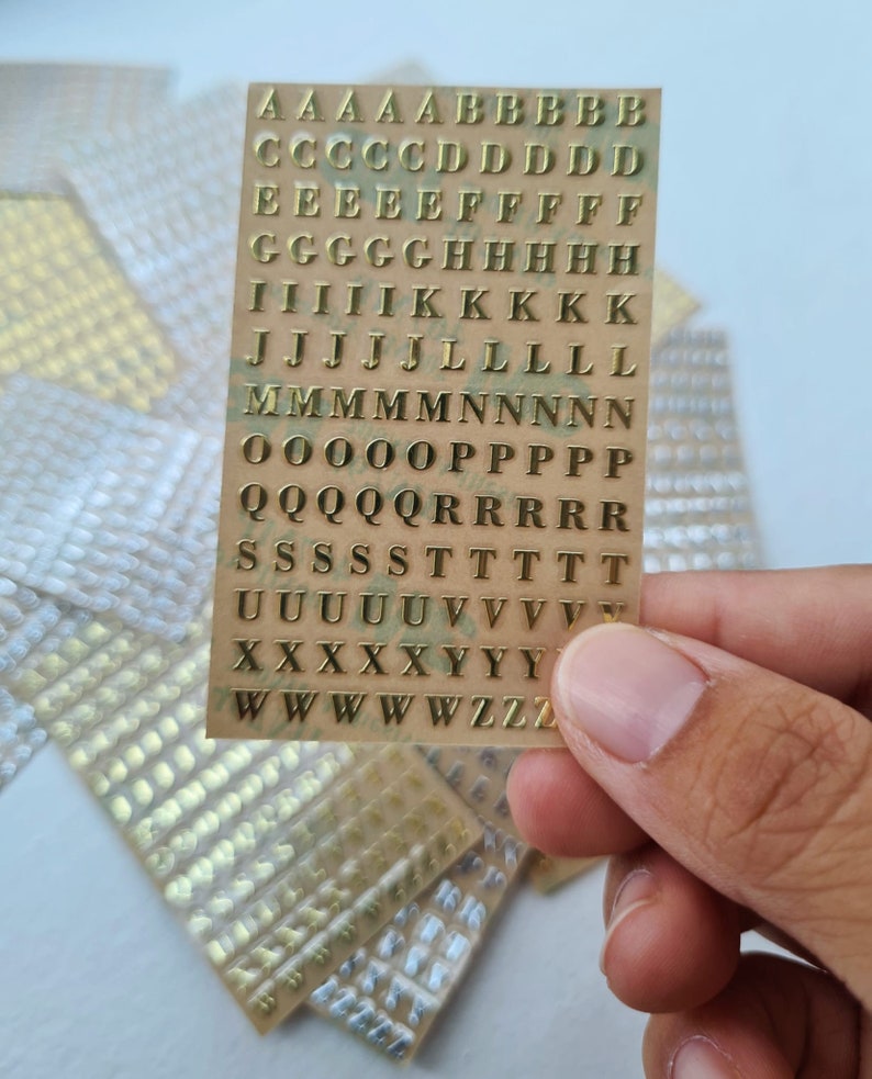 May include: A rectangular sheet of gold-coloured adhesive alphabet stickers featuring uppercase letters arranged in rows. The sheet contains multiple instances of each letter from A to Z, presented in a serif font style on a clear backing.