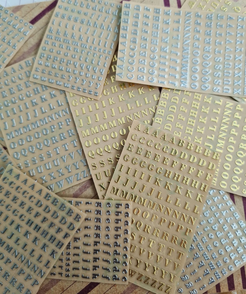 May include: Multiple rectangular sticker sheets featuring adhesive alphabet letters in both uppercase and lowercase. The letters are available in metallic silver and gold finishes, arranged in organised rows on light-coloured backing paper.