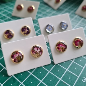 May include: Multiple pairs of round stud earrings displayed on white cards. Each earring features a gold-toned metal setting enclosing a clear resin dome containing pressed dried flowers in various colors including pink, purple, and blue.