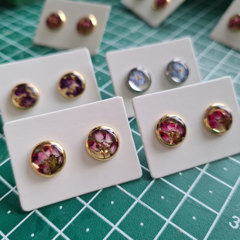 May include: Multiple pairs of round stud earrings displayed on white cards. Each earring features a gold-toned metal setting enclosing a clear resin dome containing pressed dried flowers in various colors including pink, purple, and blue.