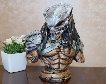 May include: A silver and gold Predator bust with a detailed sculpt of the character's head, chest, and shoulders. The bust is on a circular base.