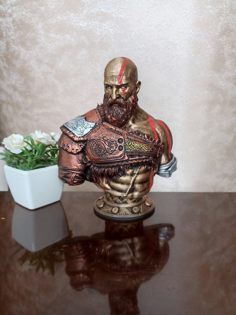 May include: A bronze bust of a bearded warrior with red markings on his face and body. He is wearing a leather and metal armor with intricate designs. The bust is on a circular base with a decorative pattern.