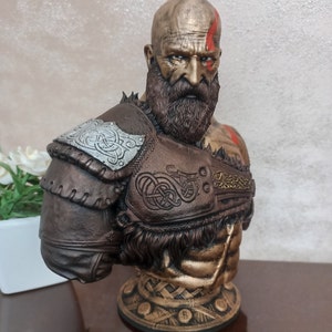 May include: A bust of a warrior with a beard and a red stripe painted on his forehead. He is wearing a brown leather armor with silver and gold accents. The bust is on a gold base.