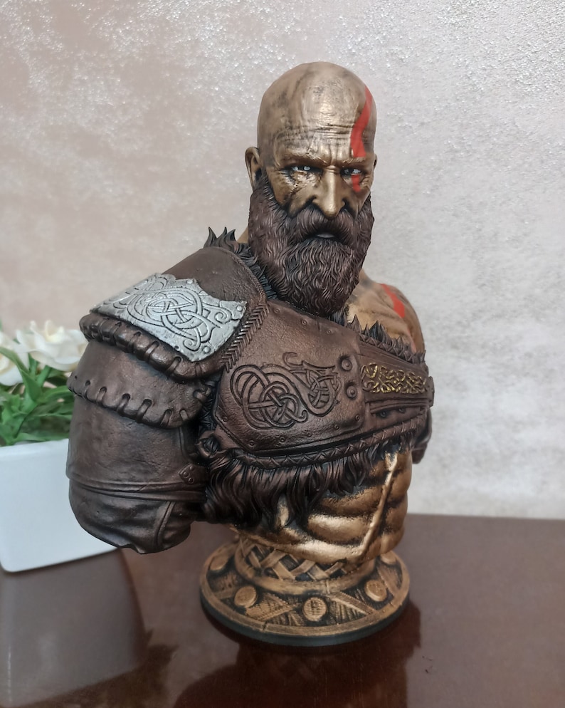 May include: A bust of a warrior with a beard and a red stripe painted on his forehead. He is wearing a brown leather armor with silver and gold accents. The bust is on a gold base.
