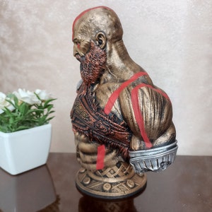 May include: A bust of a muscular figure with a red beard and red markings on the body. The figure is wearing a bronze-colored armor with intricate details. The bust is on a base with a circular design.