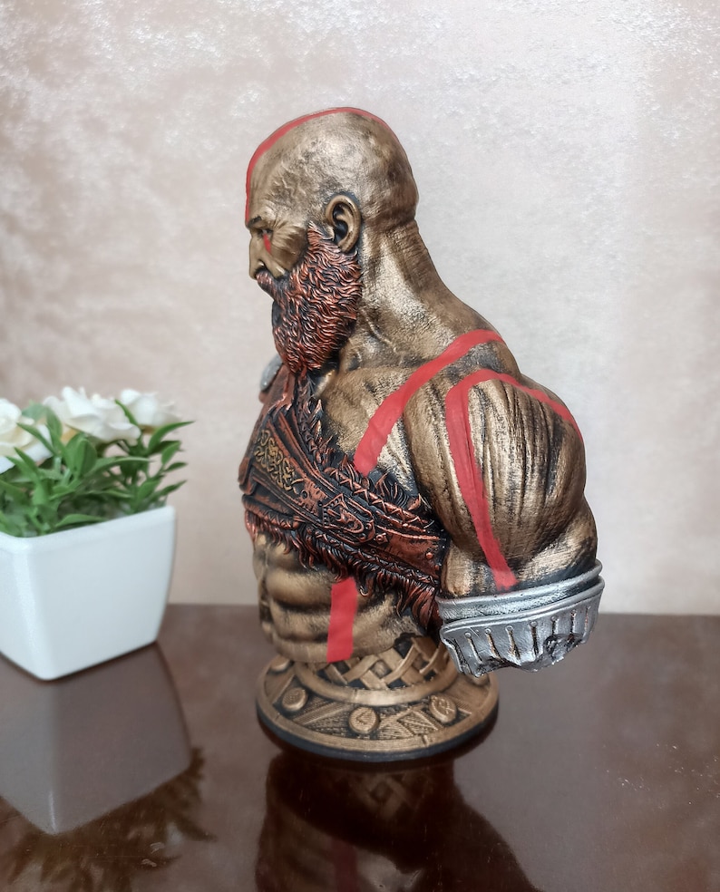 May include: A bust of a muscular figure with a red beard and red markings on the body. The figure is wearing a bronze-colored armor with intricate details. The bust is on a base with a circular design.