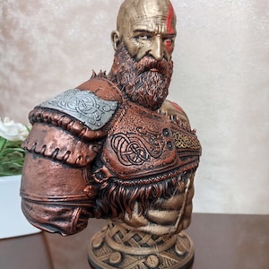 May include: A bust of a bald, bearded warrior with a red mark on his forehead. He is wearing a bronze and gold armor with intricate designs. The bust is on a bronze base with a woven pattern.