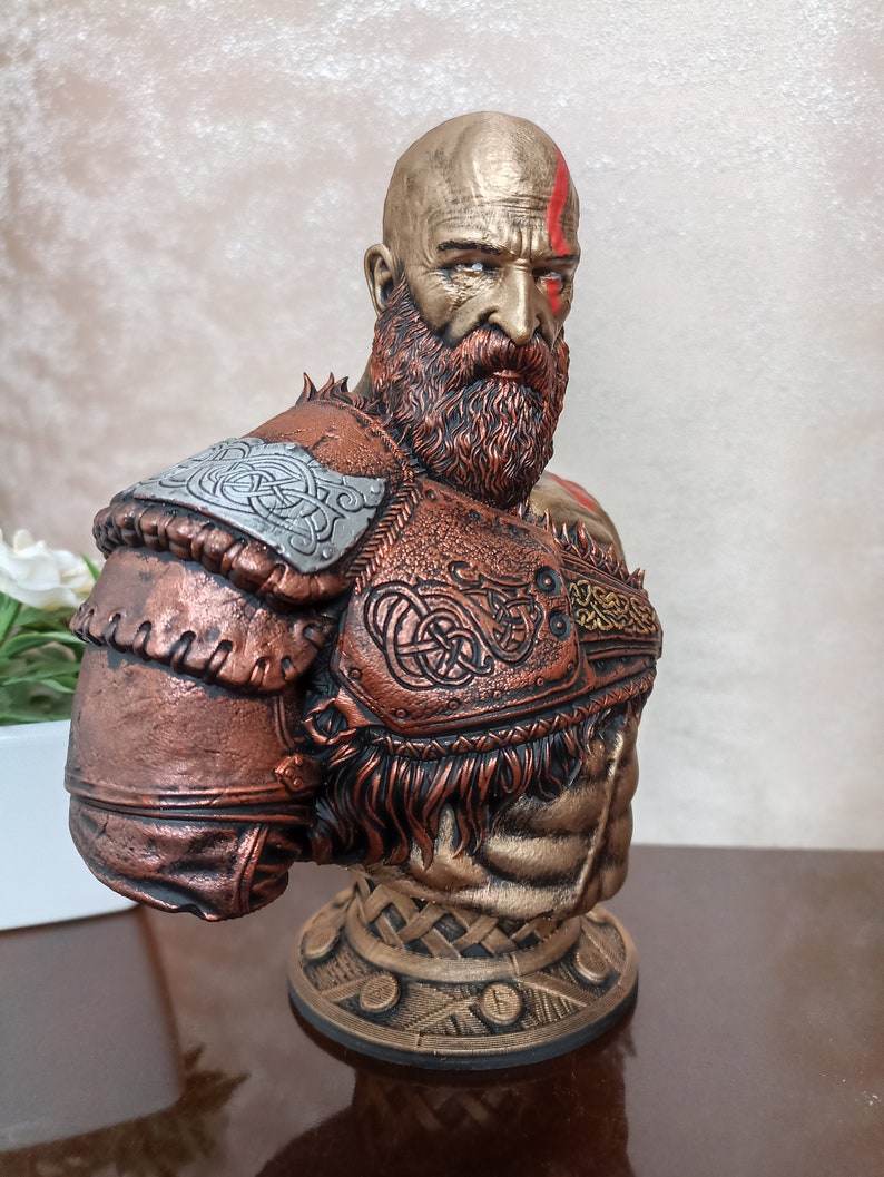 May include: A bust of a bald, bearded warrior with a red mark on his forehead. He is wearing a bronze and gold armor with intricate designs. The bust is on a bronze base with a woven pattern.