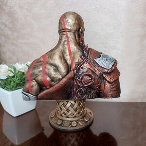May include: A bust of a warrior figure with a golden and bronze finish. The figure has red lines on its back and is wearing a brown and silver armor with a Celtic knot design. The bust is on a gold base with a woven design.