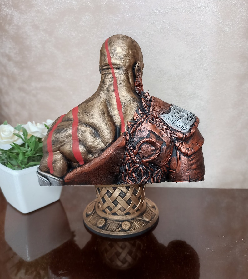 May include: A bust of a warrior figure with a golden and bronze finish. The figure has red lines on its back and is wearing a brown and silver armor with a Celtic knot design. The bust is on a gold base with a woven design.