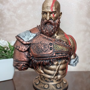 May include: A bust of a bald, bearded warrior with a red stripe across his forehead. He is wearing a bronze and gold armor with intricate designs. The bust is on a circular base with a textured design.