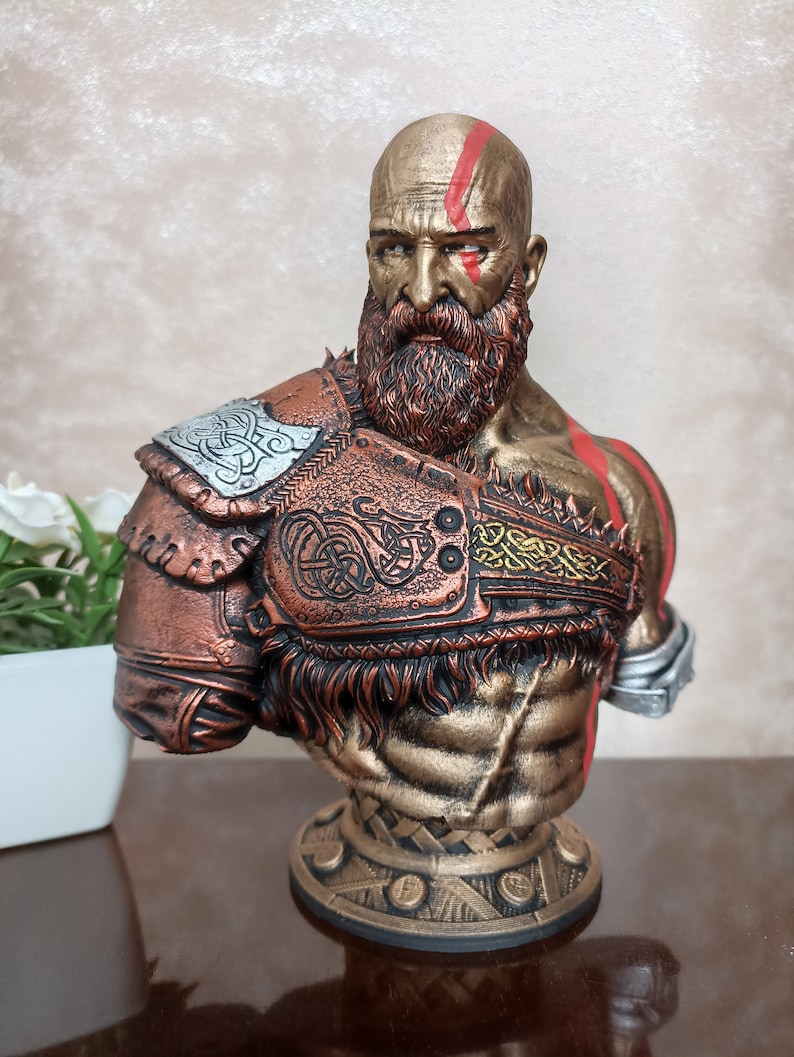 May include: A bust of a bald, bearded warrior with a red stripe across his forehead. He is wearing a bronze and gold armor with intricate designs. The bust is on a circular base with a textured design.