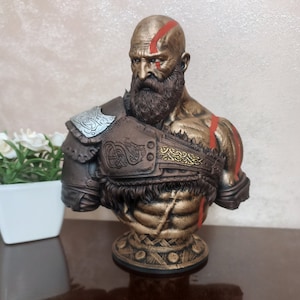 May include: A bronze bust of a bearded warrior with a red and gold design. The warrior is wearing a leather and metal armor with a braided design. The bust is on a round base with a gold and brown design.