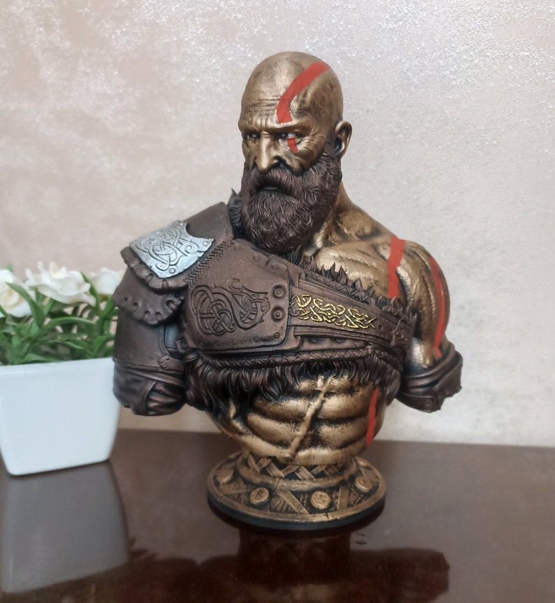 May include: A bronze bust of a bearded warrior with a red and gold design. The warrior is wearing a leather and metal armor with a braided design. The bust is on a round base with a gold and brown design.