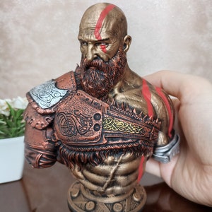 May include: A bronze bust of a warrior with a red and gold painted design. The warrior has a beard and is wearing a helmet with a braided design.