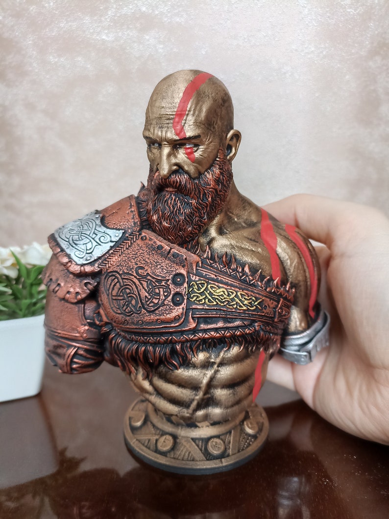 May include: A bronze bust of a warrior with a red and gold painted design. The warrior has a beard and is wearing a helmet with a braided design.