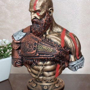 May include: A bust of Kratos from the God of War video game series. The bust is made of a gold-colored material and features red markings on the face and chest. The bust is wearing a bronze-colored armor with intricate details.