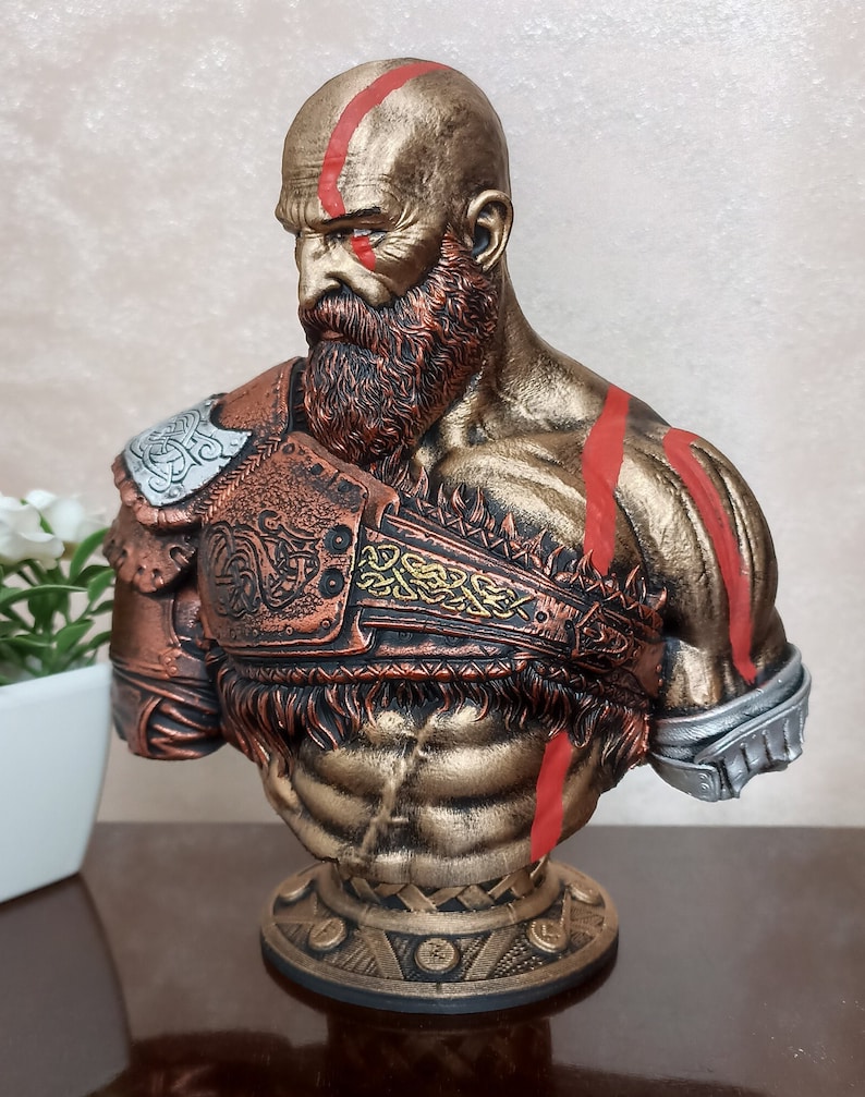May include: A bust of Kratos from the God of War video game series. The bust is made of a gold-colored material and features red markings on the face and chest. The bust is wearing a bronze-colored armor with intricate details.