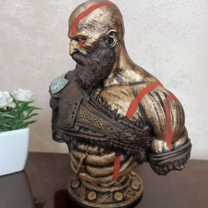 May include: A bronze bust of a bald, bearded man with red markings on his face and body. He is wearing a brown leather armor with gold accents. The bust is on a round base with a gold and black design.