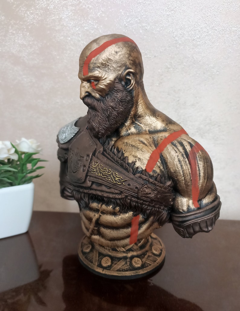 May include: A bronze bust of a bald, bearded man with red markings on his face and body. He is wearing a brown leather armor with gold accents. The bust is on a round base with a gold and black design.