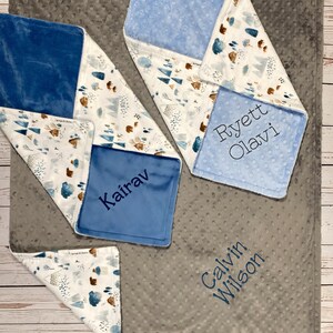 May include: Four personalized baby blankets with a gray, white, and blue color scheme. The blankets feature a woodland design with trees, mountains, and bears. The names "Kairav", "Ryett Olavi", "Calvin Wilson", and "Kairav" are embroidered on the blankets.