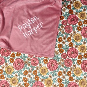 May include: A soft, dusty rose-colored blanket featuring the embroidered white text "Payton Harper" in a cursive script, partially covering a white fabric base printed with a repeating pattern of pink roses, yellow sunflowers, and teal leaves.