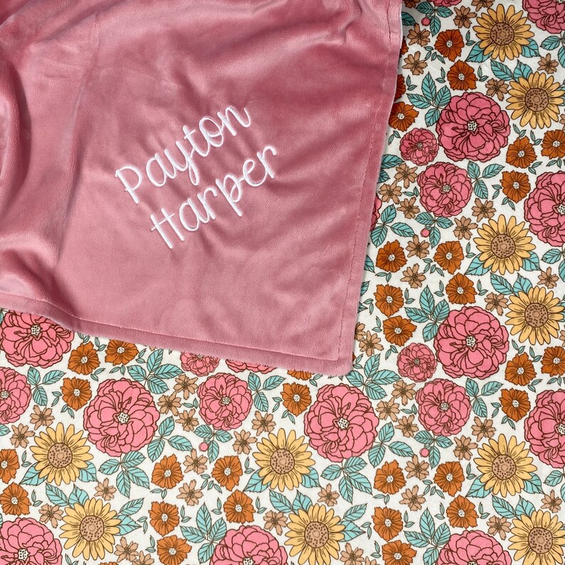May include: A soft, dusty rose-colored blanket featuring the embroidered white text "Payton Harper" in a cursive script, partially covering a white fabric base printed with a repeating pattern of pink roses, yellow sunflowers, and teal leaves.