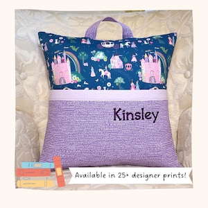 May include: A purple and blue reading pillow with a handle. The top section features a whimsical castle and rainbow pattern. The bottom section is a dotted purple fabric with the name "Kinsley" embroidered. The text "Available in 25+ designer prints!" is also visible.