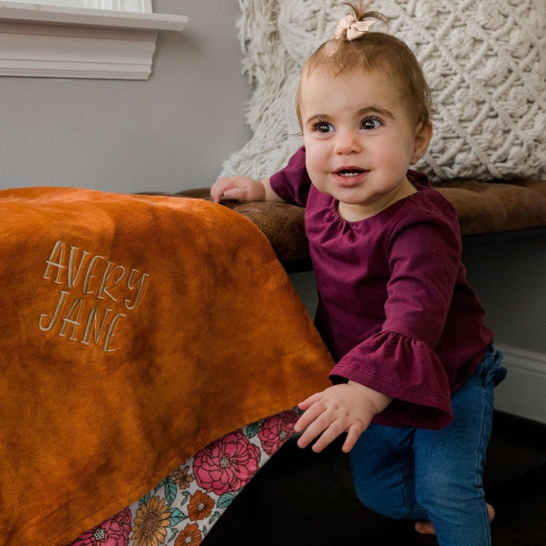 May include: A soft, burnt orange plush blanket featuring the embroidered name AVERY JANE in a light tan thread. The blanket has a decorative border with a colorful floral pattern of pink, orange, and yellow flowers on a light teal background.