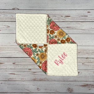 May include: A square baby security blanket featuring a cream-colored dimpled minky fabric on one side and a floral print on the other. The floral pattern includes pink peonies, yellow sunflowers, and green leaves. The name Rylee is embroidered in pink.