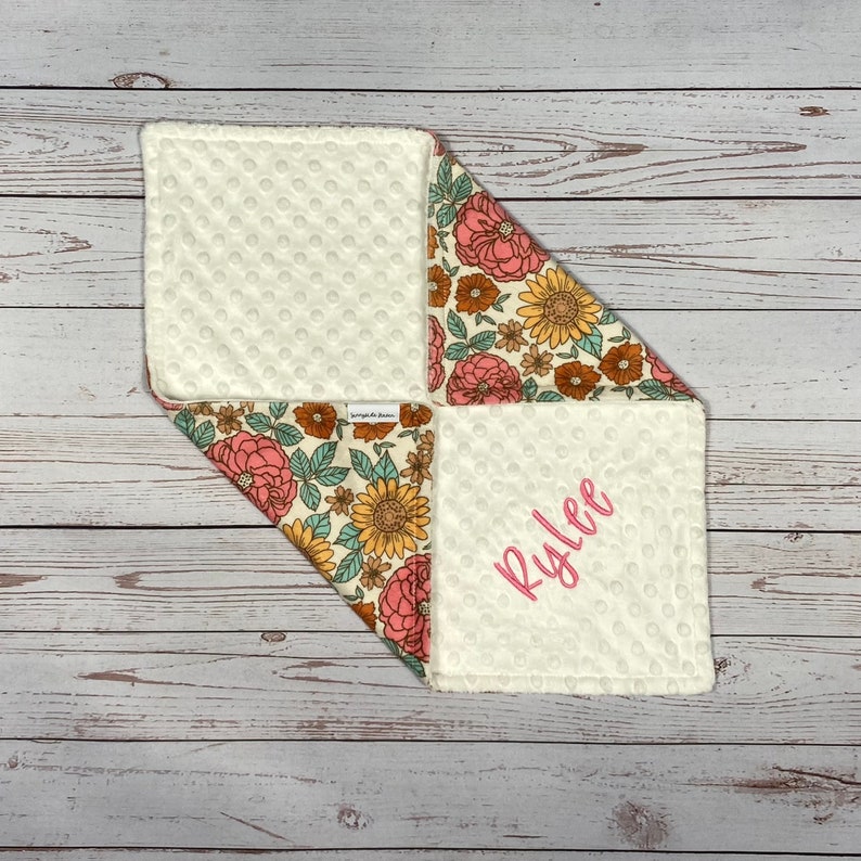May include: A square baby security blanket featuring a cream-colored dimpled minky fabric on one side and a floral print on the other. The floral pattern includes pink peonies, yellow sunflowers, and green leaves. The name Rylee is embroidered in pink.