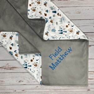 May include: A gray and white baby blanket with a woodland pattern and the name "Field Matthew" embroidered on it.