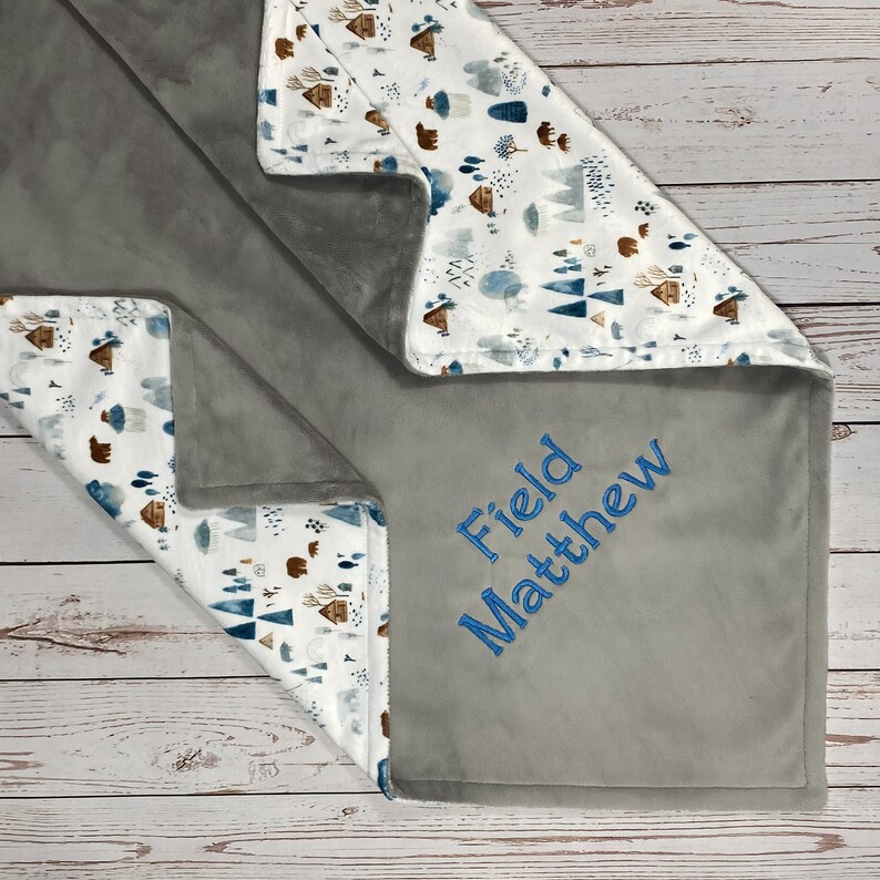 May include: A gray and white baby blanket with a woodland pattern and the name "Field Matthew" embroidered on it.
