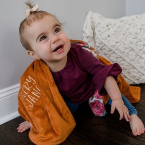 May include: A soft, burnt orange plush blanket featuring the embroidered name Avery Jane in white script lettering, draped over a toddler wearing a dark purple long-sleeved shirt and blue leggings with a floral pattern on the sleeve.