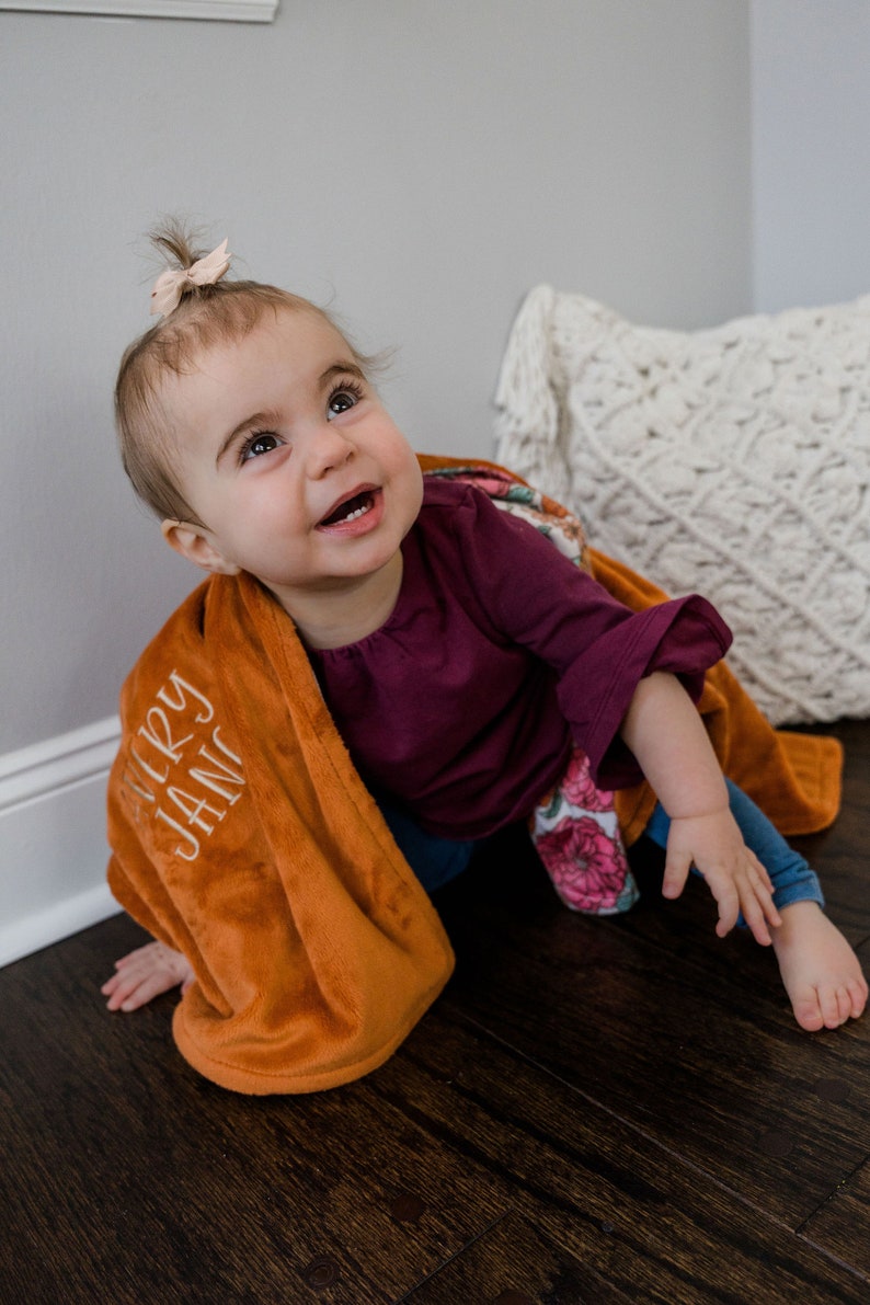 May include: A soft, burnt orange plush blanket featuring the embroidered name Avery Jane in white script lettering, draped over a toddler wearing a dark purple long-sleeved shirt and blue leggings with a floral pattern on the sleeve.