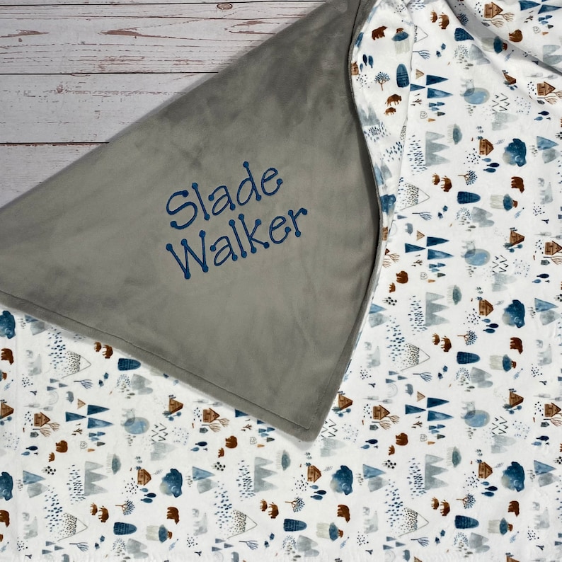 May include: A gray and white baby blanket with a blue and brown woodland pattern. The blanket has a gray side with the name "Slade Walker" embroidered in blue.
