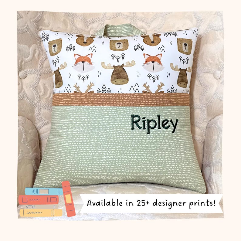 May include: A square throw pillow featuring a top section with a white background printed with brown bears, orange foxes, and moose, and a bottom section in sage green with white dots. The name Ripley is embroidered in dark green on the green section.