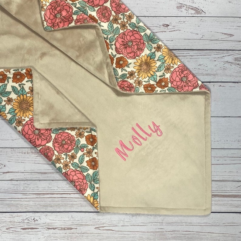 May include: A soft, plush blanket featuring a beige side embroidered with the name Molly in pink script and a contrasting side with a floral pattern of pink peonies, yellow sunflowers, and small brown flowers on a cream background.
