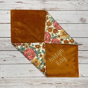 May include: A square baby security blanket featuring a soft, solid mustard-brown plush fabric on one side and a cream-colored cotton fabric with a floral pattern of pink, yellow, and brown flowers on the other. The name Ashtyn Ray is embroidered in gold thread.