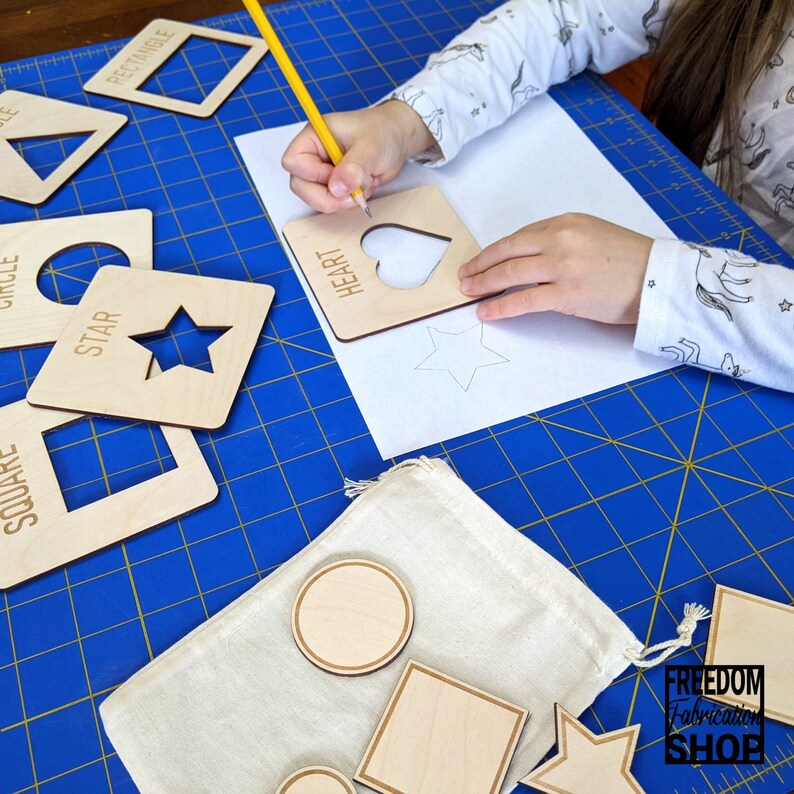 May include: A child traces shapes on a white sheet of paper using a wooden stencil set. The stencils are made of wood and have cutouts of different shapes, including a heart, star, circle, square, and rectangle. The stencils are labeled with the name of the shape. The child is using a yellow pencil to trace the shapes. The child is sitting at a table with a blue grid pattern. The table has a white bag with wooden shapes in it. The bag has a tag that says "FREEDOM Fabrication SHOP".