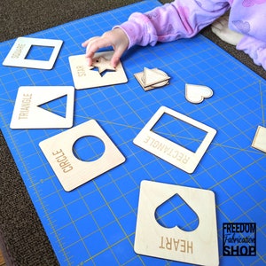 May include: A set of wooden shape puzzle pieces with cutouts of different shapes, including a square, triangle, circle, rectangle, heart, and star. The pieces are on a blue cutting mat with a grid pattern. The text "FREEDOM Fabrication SHOP" is visible in the bottom right corner of the image.