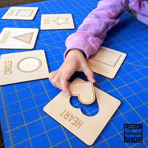May include: A child's hand holds a wooden heart shape puzzle piece over a wooden board with a heart-shaped cutout. The board is labeled "HEART". Other wooden puzzle pieces are visible in the background, labeled "SQUARE", "STAR", "TRIANGLE", "CIRCLE", and "RECTANGLE".