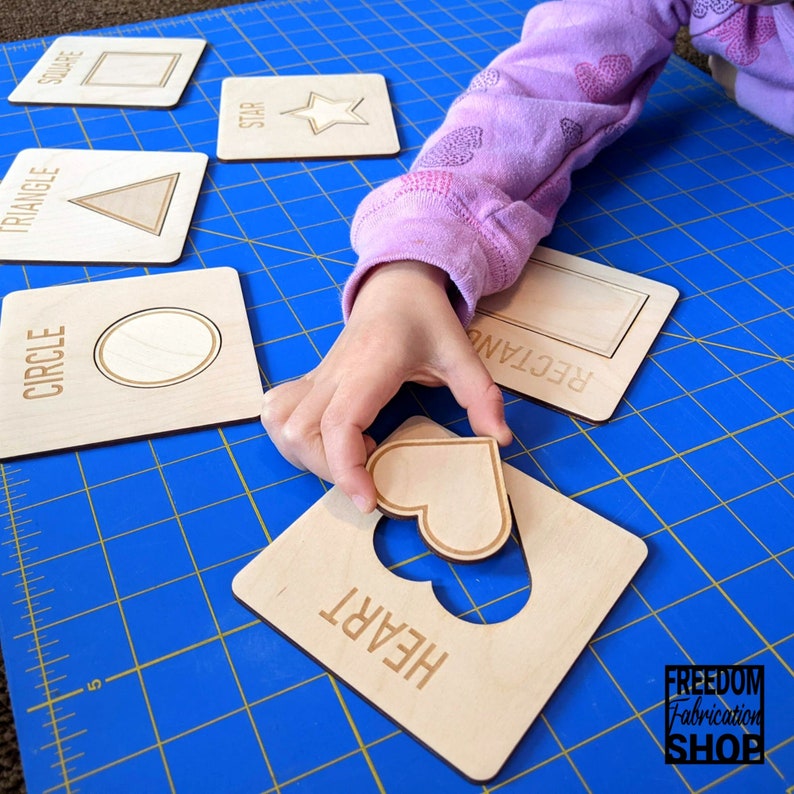 May include: A child's hand holds a wooden heart shape puzzle piece over a wooden board with a heart-shaped cutout. The board is labeled "HEART". Other wooden puzzle pieces are visible in the background, labeled "SQUARE", "STAR", "TRIANGLE", "CIRCLE", and "RECTANGLE".