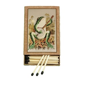 May include: A vintage matchbox with a green frog playing a banjo on the front. The matchbox is open and the matches are visible.