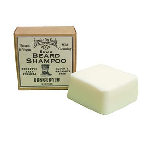 Puede incluir: Una pastilla de champú sólido e inodoro para barba junto a su caja de cartón. La caja presenta el texto "Superior Dry Goods" y "Solid Beard Shampoo". La pastilla es de 56 gramos y sin color ni fragancia.