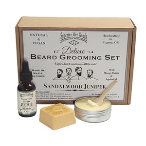 May include: A brown cardboard box with a white label that says "Superior Dry Goods Deluxe Beard Grooming Set" and "Tames And Conditions All Beards". The box contains a set of beard care products, including a bottle of beard oil, a tin of beard balm, and a bar of beard shampoo. The products are labeled "Sandalwood Juniper" and are made with mango butter and jojoba oil.