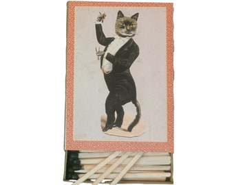 May include: A vintage matchbox with a black and white illustration of a cat wearing a tuxedo and holding a conductor's baton. The matchbox is red with a white and pink patterned border.