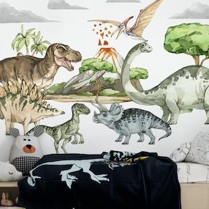 Op de afbeelding: Waterverf dinosaurus muurstickers met een Tyrannosaurus Rex, Stegosaurus en Triceratops. De scène bevat een vulkaan, bomen en een Pterodactylus. Het kleurenpalet omvat tinten groen, bruin en grijs. Een zwarte deken met een dinosaurusontwerp is zichtbaar.
