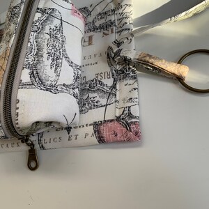 May include: A white canvas zippered pouch with a black and white print of a vintage design. The pouch has a brown zipper and a brown metal ring attached to a brown strap.